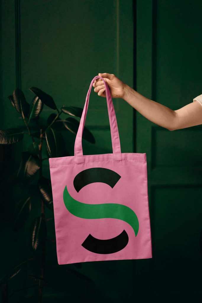 Stoffbeutel, der von einem Arm gehalten wird. Der Beutel ist rosa mit dem großen Buchstaben „S“ darauf in kräftigen Grün-Tönen.